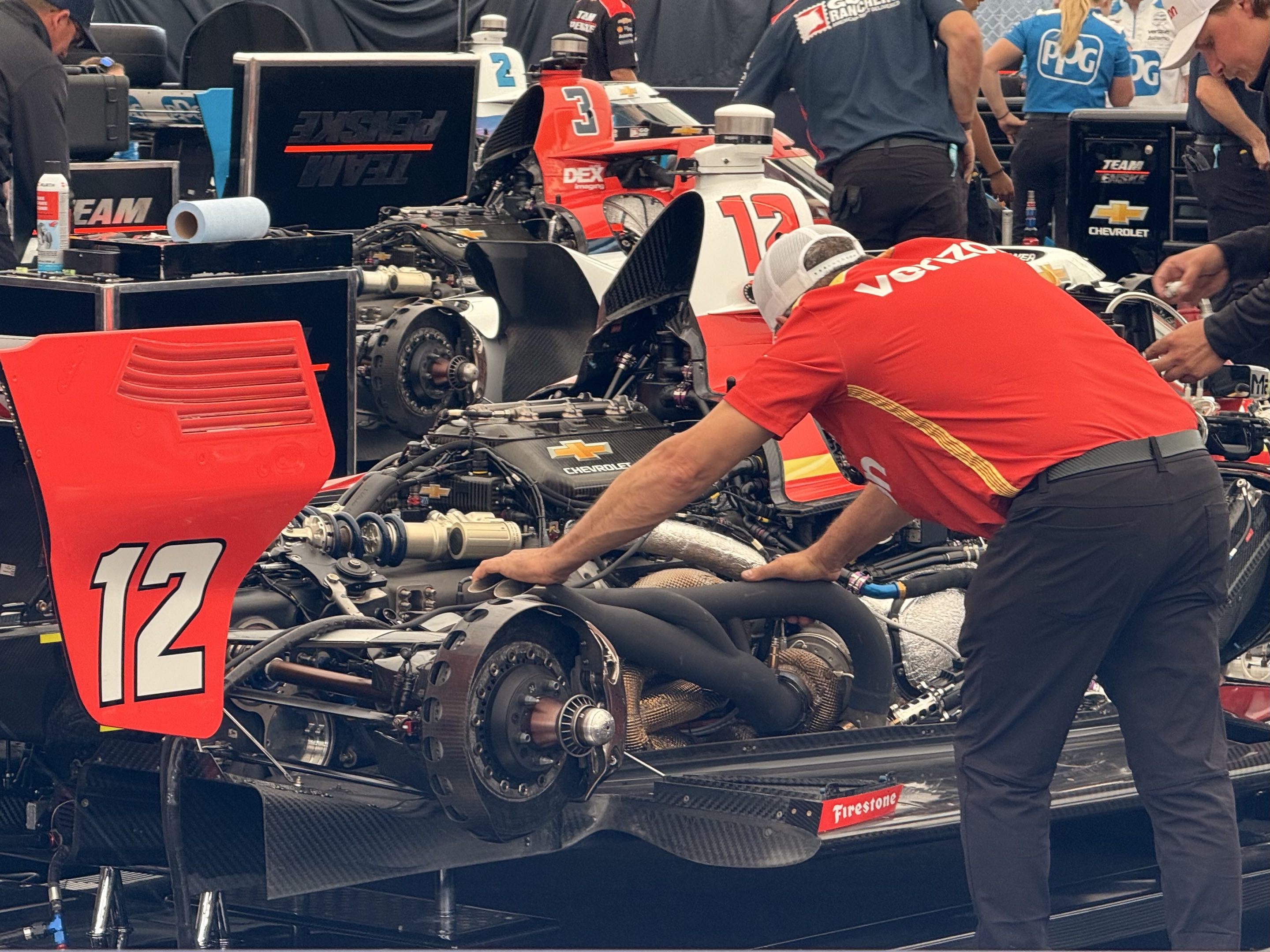 A mechanic in a Verizon polo shirt and white hat with his hands in the guts of the #12 Penske Chevrolet Indy car with its engine cover removed